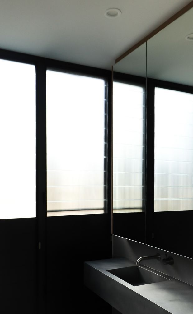 A compact, dark-toned bathroom or washroom with a wall-mounted stainless-steel or metal sink and integrated backsplash. The space features black-framed translucent windows, a large mirrored wall, a white ceiling with recessed downlights, and a minimalist monochromatic interior.