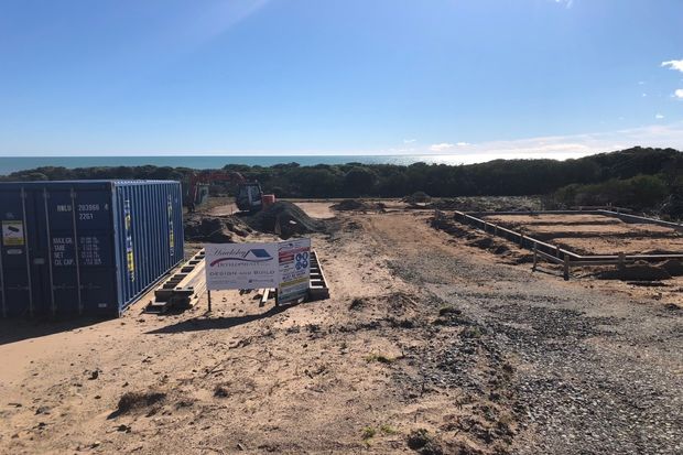 A coastal construction site viewed from an elevated position, with earthworks, timber-framed foundation sections, construction machinery, shipping containers, and a project sign. Dense coastal vegetation and the ocean are visible beyond the site.