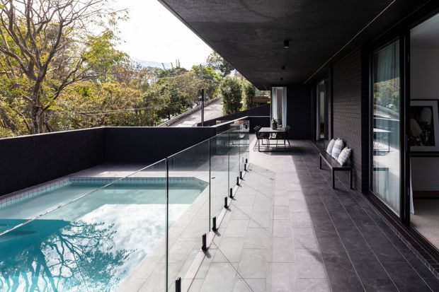 A contemporary covered outdoor terrace beside a rectangular swimming pool, enclosed by clear glass balustrades and a dark rendered wall. The terrace has large grey tiled flooring, dark brick or masonry wall cladding, full-height glazed doors, and minimalist outdoor furniture. Trees and neighbouring rooftops are visible beyond the property.