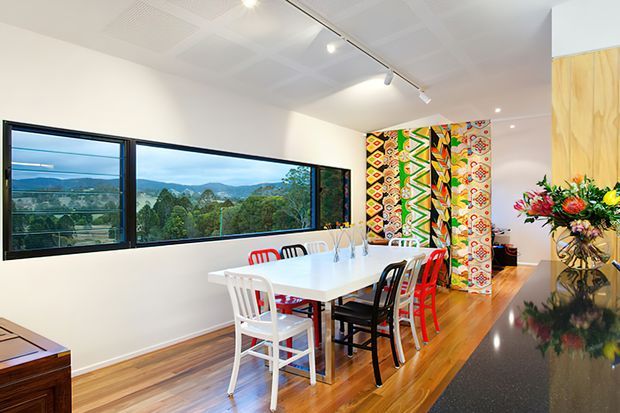 Bright open-plan dining area with a long white dining table and mismatched colourful chairs, set against large panoramic windows overlooking a green hilly landscape. The room features polished timber flooring, white walls and ceiling, a vibrant patterned textile screen, track lighting, and a dark stone kitchen benchtop in the foreground.