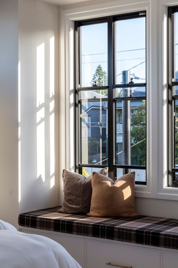 Bright bedroom corner featuring a built-in window seat beneath tall black-framed windows, with plaid upholstery, warm neutral cushions, and white built-in cabinetry. Strong sunlight casts geometric shadows across the white walls, while a suburban streetscape is visible through the glazing.