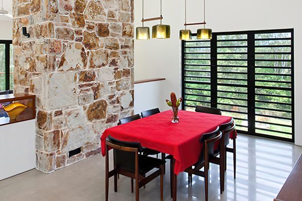 A bright dining area with a rectangular table covered in a vivid red tablecloth and surrounded by dark timber chairs. A large irregular natural stone feature wall dominates the left side, while black-framed horizontal louvred glazing opens to greenery outside. Brass and dark pendant lights hang above the table, and the polished light floor reflects the room's natural light.