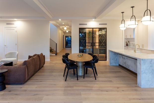 Open-plan contemporary apartment interior showing a living room, dining area and kitchen. The space features pale timber flooring, white walls and ceiling detailing, a large stone island with waterfall edge, dark cabinetry, black-framed glazed joinery, a round timber dining table, and sculptural pendant lights.
