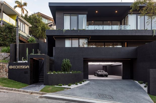 Street-facing view of a contemporary multi-level residence with a dark textured facade, flat roof, projecting upper-floor terraces, extensive glazing, integrated landscaping, and a recessed double garage. The property sits in a dense suburban setting with neighbouring homes and palm trees visible around it.