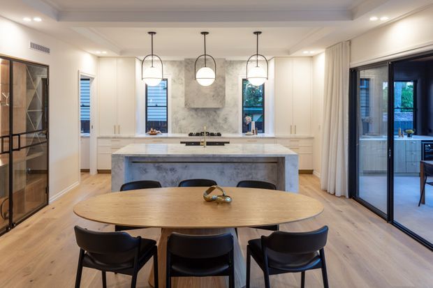 A bright, refined open-plan kitchen and dining area with a large stone island, white built-in cabinetry, timber flooring, and a curved timber dining table with black chairs. Three pendant lights hang above the island, while full-height glazed doors open to an adjoining outdoor or transitional space.