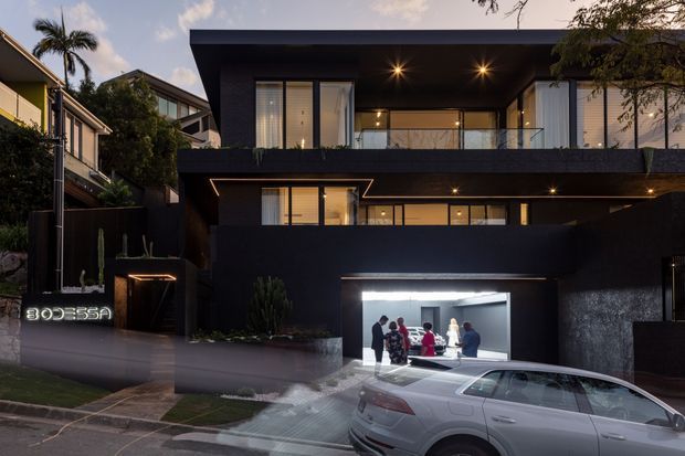 A contemporary multi-storey dark-clad residence photographed at dusk, featuring strong horizontal volumes, recessed warm lighting, upper-level glazing with glass balustrades, and an illuminated garage or showroom-like opening at street level. A vehicle is parked in front, with neighbouring homes and established landscaping visible around the property.