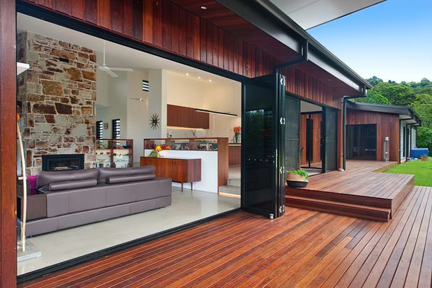 Contemporary timber-clad home with expansive black-framed glazing opening onto a broad timber deck. The open-plan interior combines a living room with a grey sectional sofa and stone fireplace, and a modern kitchen with a timber island. The deck steps down toward a landscaped garden, with lush greenery visible beyond.
