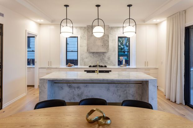 A refined contemporary kitchen viewed from the dining area, featuring a large pale stone island with integrated seating, white cabinetry, marble-look splashback and rangehood surround, timber flooring, and three sculptural globe pendant lights. Tall windows and full-height curtains provide natural light, while black-framed glazing and dark dining chairs add contrast.