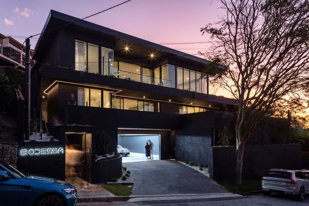 A contemporary multi-storey dark-clad urban residence photographed at dusk. The home features a dramatic angular roofline, extensive floor-to-ceiling glazing, cantilevered upper-level forms, warm recessed exterior lighting, a wide driveway, and an illuminated integrated garage. Mature trees and neighbouring buildings frame the street-facing facade.