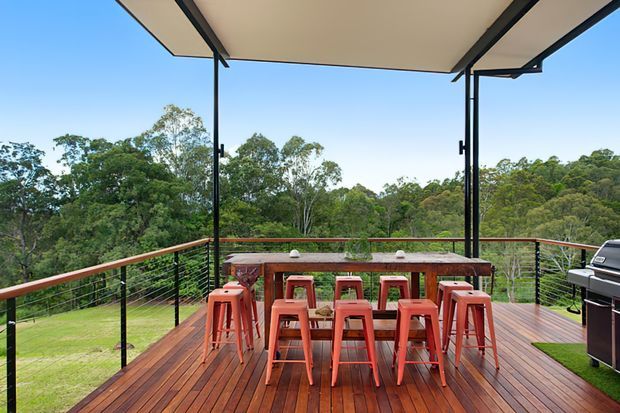 Elevated outdoor entertaining deck overlooking dense greenery, with a long timber dining table, multiple red bar stools, black cable railing, and a large retractable canopy overhead. A barbecue sits at the right edge of the deck.