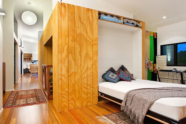 Bright compact bedroom with a built-in timber wall and fold-down Murphy bed, open shelving, patterned cushions, a textured throw, and warm timber flooring. The room connects to a hallway and appears to include integrated storage and a small desk workspace.