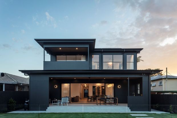 A contemporary two-storey residential house viewed from the rear at dusk. The dark-clad facade features a broad covered outdoor entertaining area, upper-level glazing, a projecting flat roof, and a small lawn enclosed by neighbouring homes and fencing.