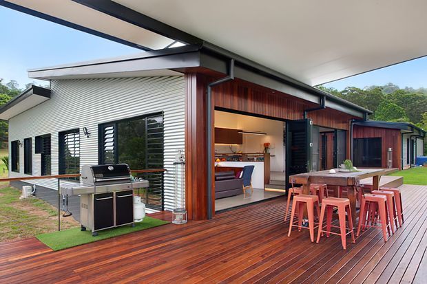 Contemporary single-storey home with a large timber deck and outdoor dining area, connected to an open-plan interior through wide openings. The exterior combines dark-stained timber cladding, light corrugated wall cladding, black-framed louvred windows, and broad roof overhangs. A built-in-style outdoor kitchen and barbecue sit beside the deck, with landscaped grounds and trees beyond.