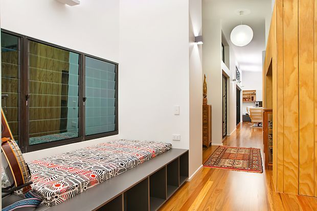 A narrow, bright interior hallway with a built-in window seat or daybed on the left, covered in a patterned textile, and extensive warm timber cabinetry along the right wall. The hallway extends toward a living area in the distance, with polished timber flooring, a patterned runner rug, modern pendant lighting, and dark-framed internal glazing.