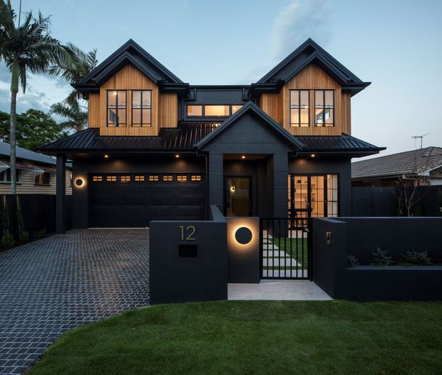 Front exterior view of a two-storey contemporary residence at dusk. The facade combines dark charcoal cladding and roofing with warm timber gable infill, prominent pitched gables, large multi-pane windows, a recessed entry, integrated garage, front gate, and patterned paved driveway.
