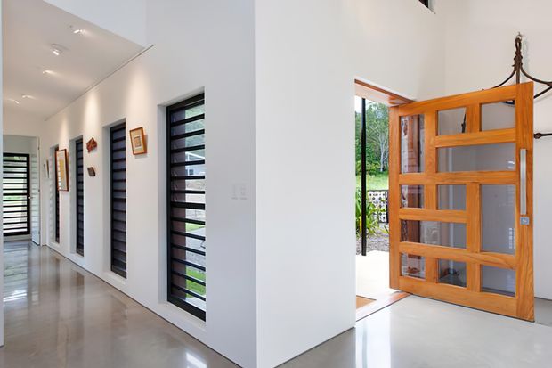 Bright contemporary residential entry hall with polished light flooring, crisp white walls, a high white ceiling with recessed downlights, and a series of narrow horizontal-louver windows along the corridor. The view looks toward a large timber-framed glazed front door with sidelights, opening to greenery outside. Small framed artworks and decorative objects line the hallway.