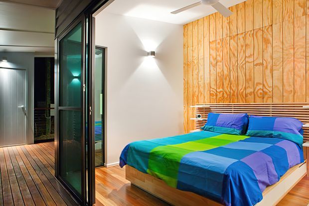 A vibrant modern bedroom with a low timber platform bed, colourful blue, green and purple bedding, and a full-height plywood feature wall behind the bed. Timber flooring continues into an adjoining glazed outdoor or circulation area, while black-framed glass doors, minimalist wall lighting, and a ceiling fan reinforce the contemporary design.
