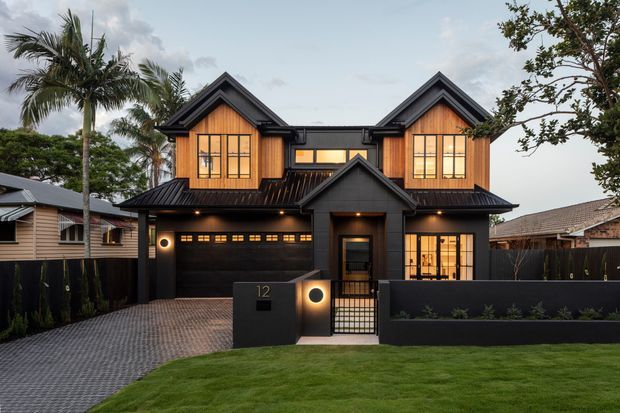Street-facing view of a contemporary two-storey residence with a symmetrical gabled facade, black metal roofing and dark cladding contrasted with warm timber-toned upper-level window bays. The home includes an integrated double garage, covered entry porch, front gate, paved driveway, low rendered boundary wall, and landscaped lawn. Palm trees and neighbouring homes are visible in the background.