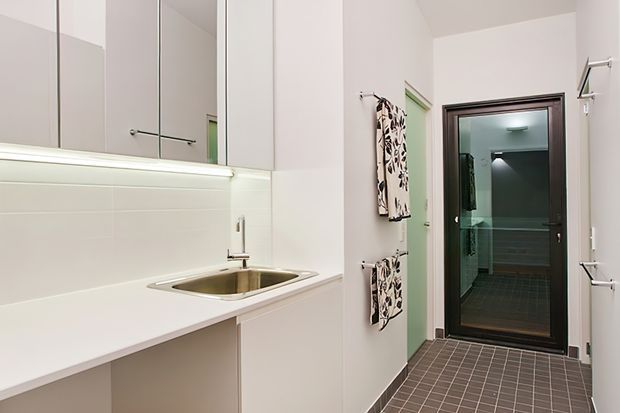 Compact, bright utility laundry with white built-in cabinetry, mirrored upper cupboards, a white benchtop, integrated stainless-steel sink, tiled splashback, and dark tiled flooring. A glass door leads to a separate tiled bathroom or wet area, with wall-mounted drying rails and patterned hanging textiles along the corridor.