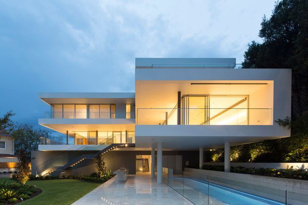Twilight exterior photograph of a large contemporary multi-level residence with crisp white rectangular volumes, extensive floor-to-ceiling glazing, projecting balconies, a flat roof, and a covered entry or carport-like area. A long narrow swimming pool and landscaped garden run alongside the polished forecourt, with warm interior and landscape lighting visible.