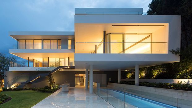 Twilight exterior view of a large contemporary multi-level residence with crisp white rendered volumes, extensive floor-to-ceiling glazing, cantilevered upper floors, a flat roof, and warm interior lighting. The home fronts a long narrow swimming pool and paved terrace, with landscaped planting and an exterior stair connecting the levels.
