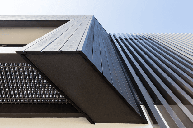 Low-angle close-up of a contemporary building facade, featuring dark vertical metal fins, a projecting angular canopy clad in grey metal panels, and a textured grid-like screen over glazing. The composition emphasises strong linear geometry, layered facade elements, and contrasting dark and light surfaces against a pale sky.