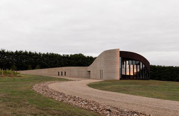 A low-profile contemporary building set within an open landscaped site, with curved sculptural walls in a pale textured finish and a dramatic rounded glazed end facade framed in dark material. A gravel driveway curves toward the entrance, with a stone drainage strip and dense hedging along the rear boundary under an overcast sky.