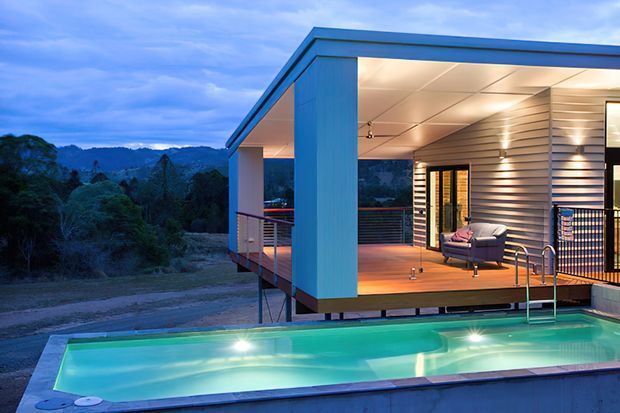 Modern elevated pavilion-style home with a flat roof, blue-grey exterior cladding, and an open covered deck overlooking a wooded landscape. A small illuminated swimming pool occupies the foreground, with contemporary outdoor living furniture, metal balustrades, and large glazed openings connecting the deck to the interior.