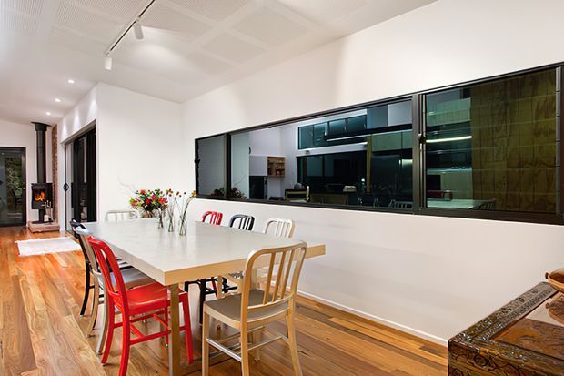 Bright open-plan dining area with a long light-coloured dining table, mismatched red, black, white and timber chairs, timber flooring, a modern tiled ceiling, and a large internal glazed partition revealing a kitchen or living space beyond. A compact freestanding fireplace is visible at the far end, with an ornate timber console in the foreground.