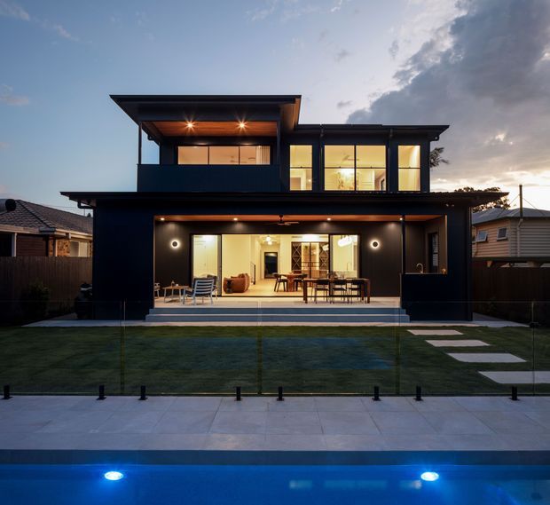 Twilight exterior view of a contemporary two-storey dark-clad residence with broad glazed openings, covered outdoor entertaining area, landscaped lawn, and a swimming pool in the foreground. The house features projecting flat roof forms, warm interior lighting, and a transparent pool fence.