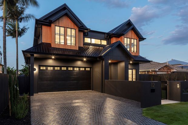 Contemporary two-storey suburban residence photographed at dusk, featuring dark metal gable roofs, warm timber-clad upper-level walls, large divided-pane windows, a black garage door, and a broad paved driveway leading to the entry. Palm trees and neighbouring homes are visible around the property.