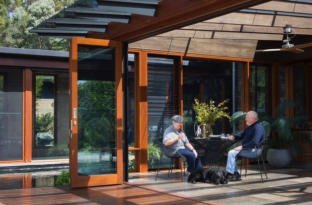 A covered outdoor terrace adjoining a modern timber-clad home, with large glazed sliding or folding doors opening into the interior. The terrace has timber decking, dark-framed glazing, exposed timber roof structure, a ceiling fan, and a small outdoor dining setting occupied by two people. A shallow reflective water feature and lush planting are visible alongside the glazed facade.