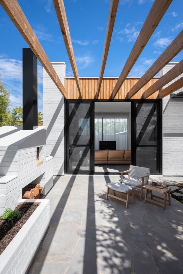 A bright contemporary courtyard patio with a white-painted brick exterior, black-framed glazed folding doors, and a timber pergola casting strong linear shadows across the stone-paved terrace. A built-in white brick outdoor fireplace with stacked firewood sits to the left, while timber-framed lounge chairs and a low table furnish the patio. Through the open doors, a living room with a tan sofa and sheer curtains is visible.