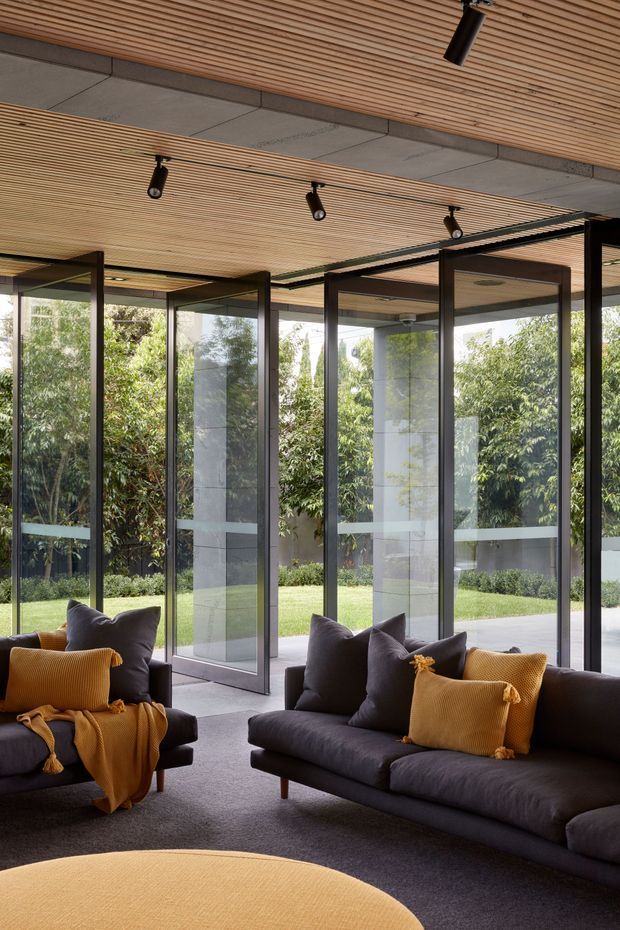 A contemporary, light-filled living room with dark upholstered sofas and armchairs arranged around a large rounded mustard ottoman. Floor-to-ceiling glazed sliding or folding doors open onto a landscaped garden, while a slatted timber ceiling with integrated black track lighting spans the room. Concrete ceiling panels and dark slim frames add a restrained modern architectural character.