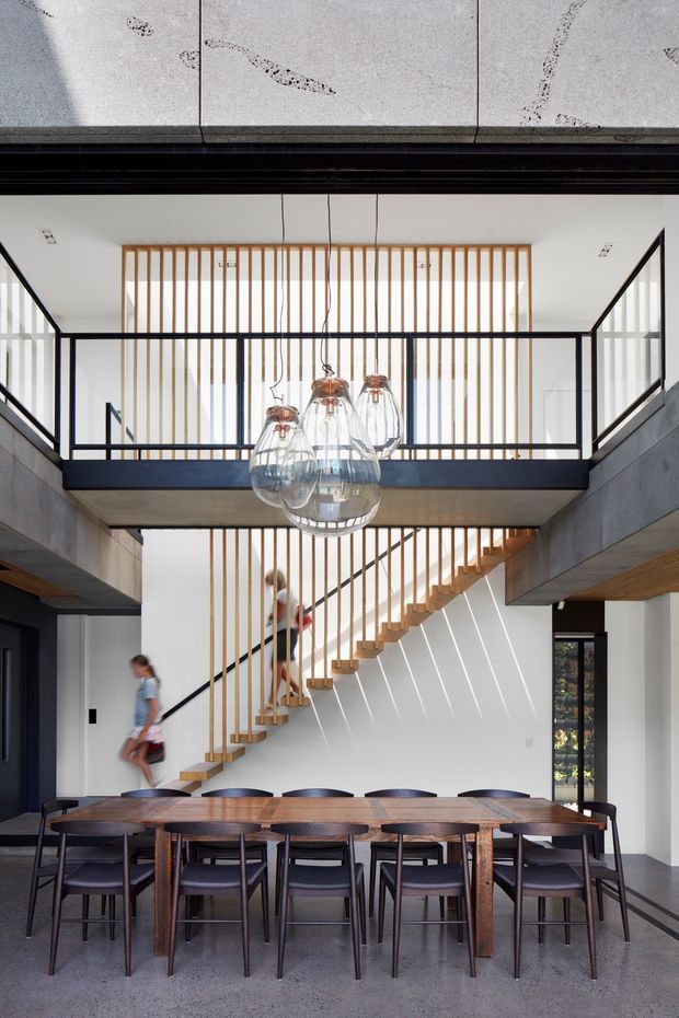 A contemporary double-height dining area with a long dark timber dining table and upholstered dining chairs in the foreground. An open-riser timber staircase climbs behind the table, enclosed by vertical timber battens and bordered by black metal-framed mezzanine balustrades. A cluster of clear glass pendant lights hangs centrally over the dining space. Concrete-toned structural elements, polished grey flooring, white walls, and extensive black-framed glazing create a refined industrial-modern interior.