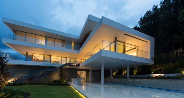 Twilight exterior view of a large multi-level contemporary residence with sharp cantilevered volumes, extensive floor-to-ceiling glazing, broad balconies, and a covered outdoor terrace. Warm interior lighting contrasts with the cool blue evening sky, while a long reflective pool or water feature runs alongside the house and landscaped garden areas surround it.