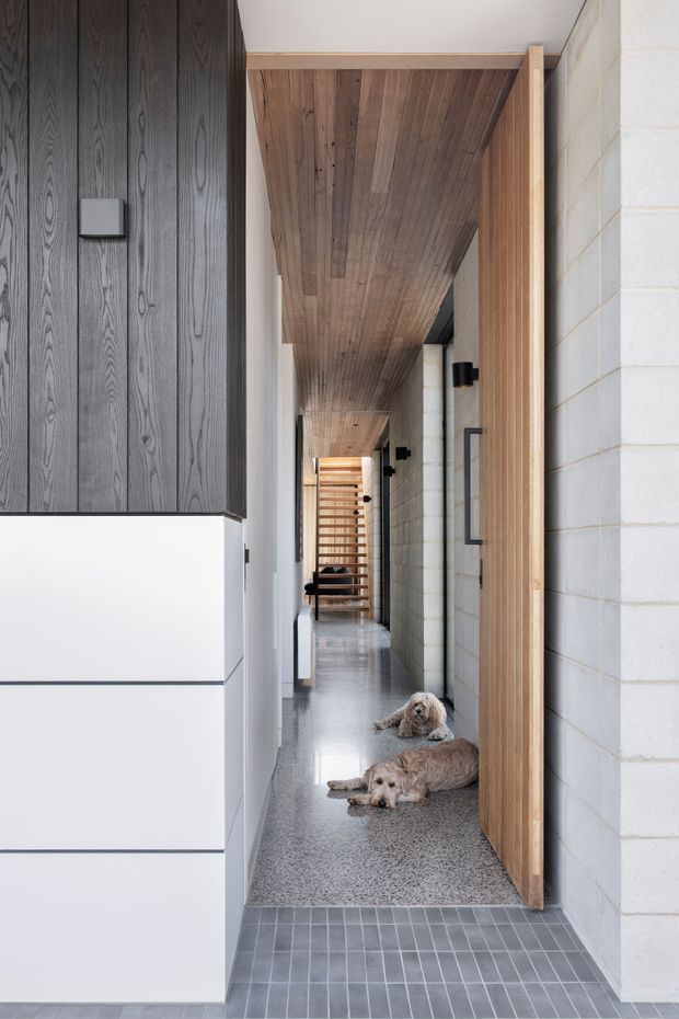A narrow modern residential hallway with a polished grey terrazzo floor, white-painted walls, a dark vertical timber-panelled built-in section, and a white painted concrete-block wall. The ceiling is lined with warm timber boards extending down the corridor, complemented by a tall timber door or screen frame on the right. Black wall sconces and a timber slatted screen are visible toward the end of the passage.