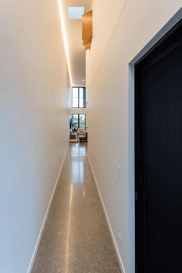 A long, narrow interior hallway with very tall white walls and a high ceiling. The polished speckled floor reflects a linear recessed light running along the ceiling, while a dark-framed doorway dominates the right foreground. Timber wall-mounted feature panels appear near the upper end of the corridor, with a bright living area visible beyond.