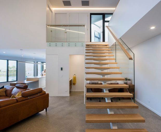 Bright contemporary open-plan interior centred on a floating timber staircase with a slim metal structure and glass balustrade. The stair rises beside a double-height void and overlooks a living area with a brown leather sofa, while a glazed upper landing and large windows bring in natural light. White walls and ceiling, recessed lighting, and polished concrete-style flooring create a clean modern feel.