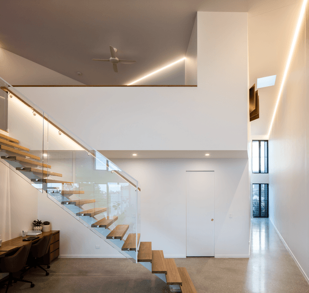 Bright contemporary double-height interior featuring a floating timber staircase with glass balustrades, polished concrete flooring, a compact work area beneath the stairs, tall vertical windows, concealed doors, and integrated linear lighting. A ceiling fan and upper-level void emphasize the open multi-level layout.