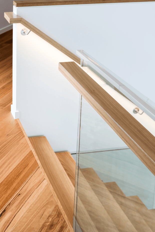 Close-up architectural view of a contemporary stairway with warm timber treads and handrail, clear glass balustrades, and crisp white walls. The stair design is light, minimal, and open, with timber flooring continuing through the landing and reflections visible in the glazing.