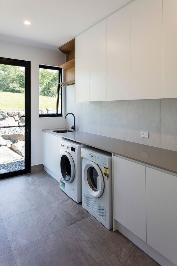A bright, contemporary laundry room with a long grey benchtop, integrated sink and black tap, front-loading washing machine and dryer, extensive white overhead and base cabinetry, and open timber shelving. Large black-framed glazing and a glazed exterior door bring in natural light and overlook a landscaped rocky garden. Large-format grey floor tiles complete the space.