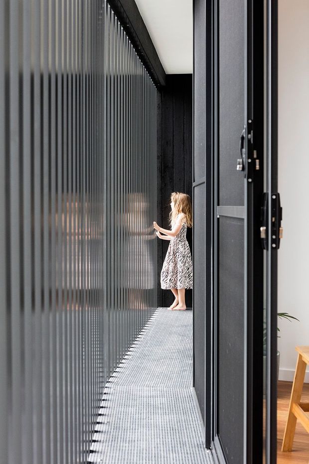 A narrow elevated walkway or balcony runs alongside a dark vertical slatted screen and full-height black-framed glazing. A person stands at the far end, while part of a warm timber-floored interior with a wooden chair is visible through the open doorway. The image emphasizes the dramatic linear enclosure, translucent privacy screen, and monochrome architectural detailing.