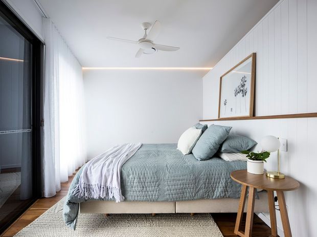 A bright, compact contemporary bedroom with a neatly made bed in soft grey-blue bedding, layered pillows, and a draped throw. The room features white vertical wall panelling with a timber picture ledge, framed botanical artwork, sheer curtains over full-height glazing, warm timber flooring, a woven rug, and a small timber bedside table with a modern lamp and potted plant. A ceiling fan and concealed warm cove lighting add practical and atmospheric details.