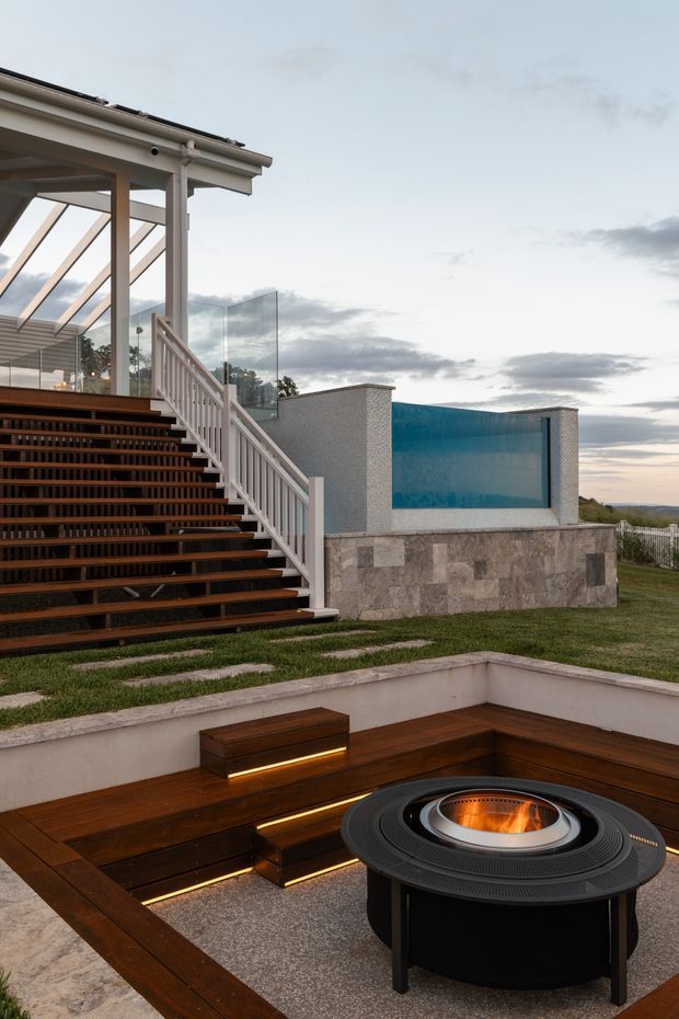 A contemporary outdoor entertaining area beside a home, photographed at dusk. The foreground features a built-in timber bench with integrated warm lighting around a circular fire pit on a paved terrace. Behind it are broad timber stairs with white balustrades leading to a glazed, covered outdoor room or sunroom. A raised rectangular swimming pool with blue-tiled or painted interior sits alongside the house, with lawn and open sky beyond.