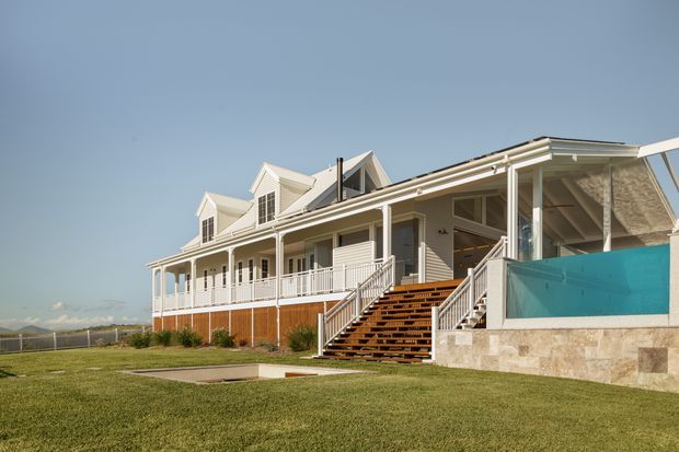 Exterior view of a large coastal-style multi-level residence with pale weatherboard cladding, multiple gabled roof forms, extensive white verandah railings and posts, and broad timber stairs leading to the raised entry. A turquoise swimming pool is visible at the right, with open lawn and distant landscape surrounding the house.