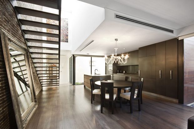 Open-plan contemporary dining and kitchen interior with a dramatic floating or open-riser staircase along a dark brick wall. The space features dark timber-look flooring, extensive glazing, a dark built-in cabinetry wall, a dining table with chairs, a compact kitchen island, white ceilings, and a sculptural chandelier. A large mirror reflects the staircase and adds depth to the room.