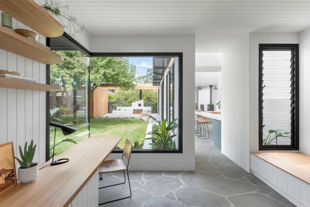 Bright contemporary open-plan interior with a built-in timber desk and open shelving along the left wall, overlooking a landscaped courtyard through large black-framed glazing. A kitchen and dining area are visible through the opening at right. The room features white vertical wall panelling, a white lined ceiling, dark irregular stone flooring, a built-in window seat, and abundant indoor planting.