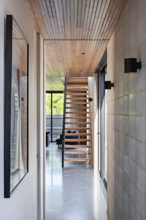 A narrow, bright interior hallway leading to an open timber staircase. The corridor features a polished grey concrete floor, exposed grey concrete block walls, a full-length timber-lined ceiling, black wall sconces, dark-framed glazing, and a large framed artwork or mirror on the left. A living area is partially visible beyond the stairs, with abundant natural light and greenery outside.