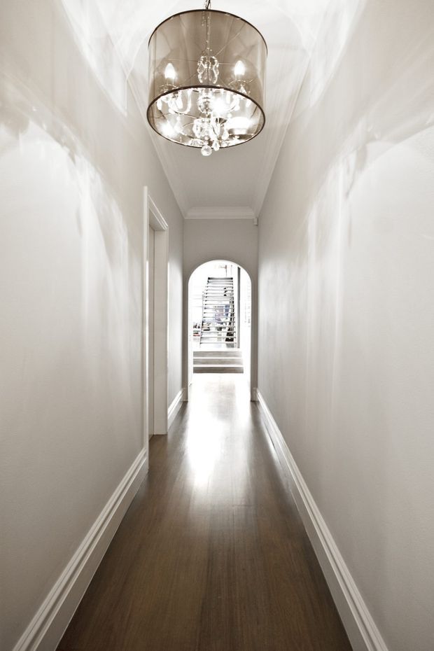 A narrow, bright residential hallway with white painted walls, decorative skirting boards and ceiling cornices, and a dark timber-look floor. A large cylindrical pendant chandelier with crystal detailing hangs centrally, while an arched opening at the far end leads toward a brighter stair or entry area with horizontal blinds.